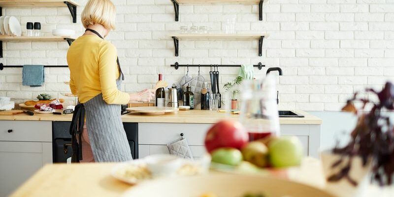 Focus on blond-haired woman in stripped apron standing at kitchen counter and cooking food for dinner